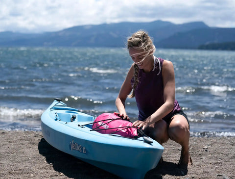 Floating Waterproof Dry Bag Backpack 10L Pink For Kayaking Rafting Boating And Outdoor Adventures next to a blue kayak on the shore