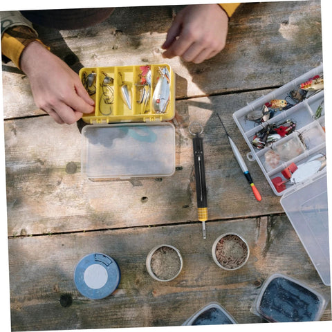 Front view of KICHOUSE stainless steel detacher tool beside a yellow tackle box with hooks and lures on a wooden table