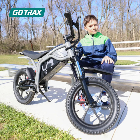 Off Road Electric Dirt Bike With 16 Inch Pneumatic Tires 300W Brushless Motor 15.5 MPH Top Speed 16 Mile Range Dual Suspension And Disc Brake Trail Ready Ride For 47 To 67 Inches Black showcased near bleachers outdoors with a young rider sitting beside.