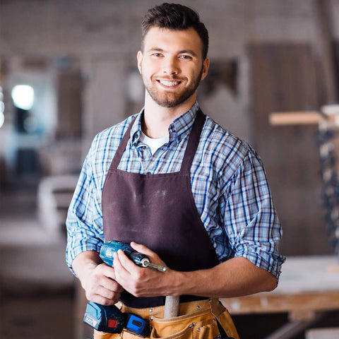 Man in workshop holding a power tool, wearing an apron and smiling, suitable for woodworking or metalworking projects.