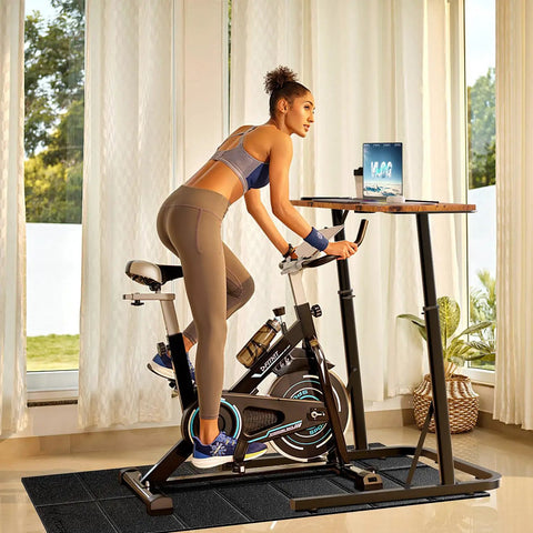 Person using adjustable standing desk for treadmills and bike workouts with laptop on a stationary bike in a home gym setup.