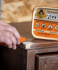 Gorilla Wood Filler 16 oz tub, natural finish; hand applying white filler to dark wood edge with a putty knife.
