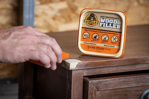 Gorilla Wood Filler 16 oz tub, natural finish; hand applying white filler to dark wood edge with a putty knife.