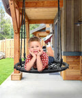 Child enjoying Saucer Tree Swing For Kids, 24 Inch Net Seat Disc Round Swing, on porch with wooden beams and greenery in background.