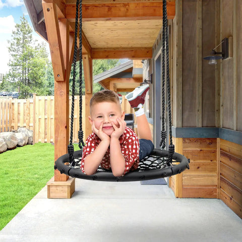 Child enjoying Saucer Tree Swing For Kids, 24 Inch Net Seat Disc Round Swing, on porch with wooden beams and greenery in background.