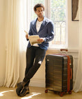 Man reading book next to 3 piece polycarbonate luggage set with black hardshell design in bright room.