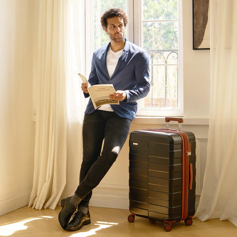 Man reading book next to 3 piece polycarbonate luggage set with black hardshell design in bright room.