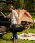 Man carrying Portable Folding Camping Table With Carry Bag And Detachable Storage Pockets at a campsite