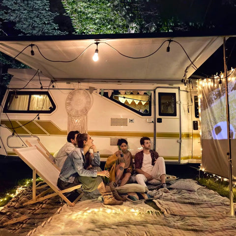 Travel trailer camping scene: white camper with gold stripe, group of friends on blankets under string lights.