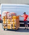 Man standing beside van with cart stacked with boxes and packages outdoors.