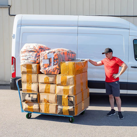 Man standing beside van with cart stacked with boxes and packages outdoors.