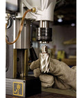 Close-up of magnetic drill press chuck with a gloved hand holding a spiral drill bit over a metal workpiece.