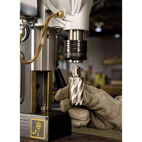 Close-up of magnetic drill press chuck with a gloved hand holding a spiral drill bit over a metal workpiece.