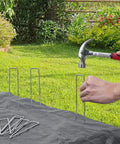 6 Inch Galvanized Landscape Staples 300 Pack U Shaped Garden Stakes Rust Proof Ground Pins For Landscaping Fabric Irrigation Tubing Fence Tent Sod Lawn Yard Outdoor Projects being hammered into grass to secure landscape fabric.