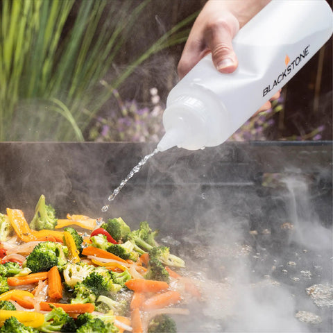 Hand using bottle to add water to vegetables on grill with Professional 22 Piece Griddle And BBQ Tool Kit Stainless Steel Dishwasher Safe For Quick Cleanup And Built To Last For Novice And Pro Grill Masters At Home And On The Grill.