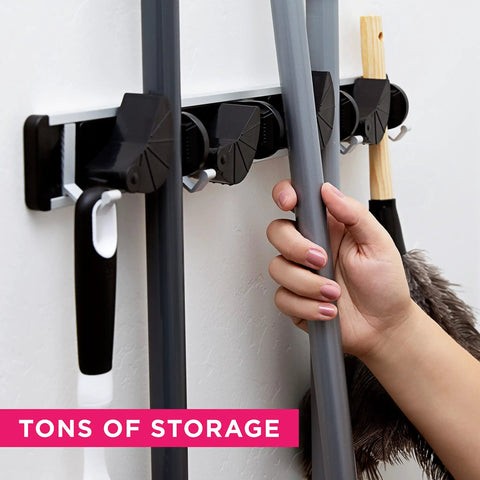 Hand placing broom in Beefy Heavy Duty Broom Wall Mount Organizer With Four Slots And Four Hooks For Brooms Mops Rakes And Garden Tools Holds Up To 1.25 Inches Handles Indoors Or Outdoors Storage For Kitchen Garage And Laundry Room