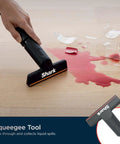 Hand using Shark squeegee tool to clean spilled liquid from a broken glass on a wooden floor.