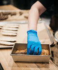 Blue nitrile disposable glove on a hand reaching into a metal baking tray with crumbs.