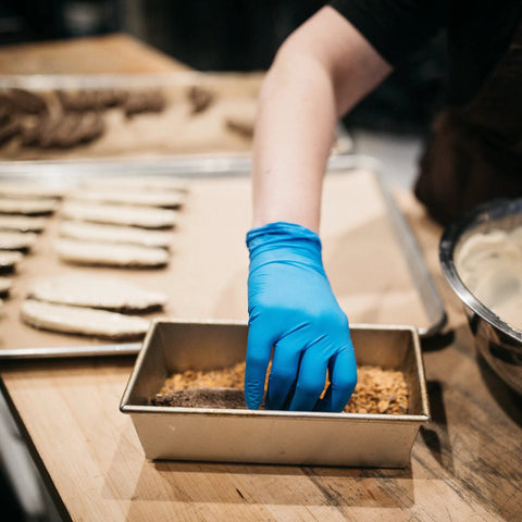 Blue nitrile disposable glove on a hand reaching into a metal baking tray with crumbs.