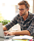 Man wearing flannel shirt and glasses using laptop at desk with headset on, focused on work