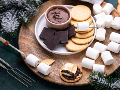 Ingredients and marshmallow roasting sticks for smores, showing chocolate, crackers, marshmallows, on wooden board.