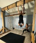 Indoor wooden fitness structure with ropes and bars, man hanging from overhead bar, woman exercising nearby, padded floor mat.