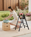 Three Step Folding Ladder With Wide Anti Slip Pedals And Comfort Handgrip Lightweight Aluminum 330 Lbs Capacity Portable Plant Support beside woman watering plants