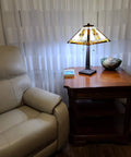 Mission Banker Style Stained Glass Table Lamp 23 Inch Tall With White Tan Brown Yellow Antique Vintage Tribal Glass For Cozy Warm Glow And Bedside Or Living Room Vibe Handmade Gift LED Bulbs Compatible on wooden table beside beige chair.
