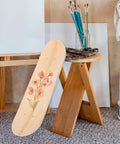 Blank Unfinished 7 Ply Wooden Skateboard Deck With Double Tail For Art Painting And Do It Yourself DIY Custom Builds Replacement Deck featuring floral art leaning against a wooden stool in an art studio.