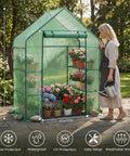 A woman waters plants inside an Outdoor Walk In Greenhouse 55 By 28 By 78 Inches With Three Tier Shelves Tool Free Setup Heavy Duty Durable PE Cover Weather Shield UV Protected For Garden And Backyard on a patio.