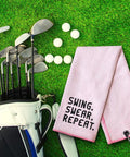 Swing Swear Repeat Embroidered Pink Golf Towel With Clip For Golf Bags next to golf clubs on grass