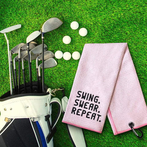 Swing Swear Repeat Embroidered Pink Golf Towel With Clip For Golf Bags next to golf clubs on grass
