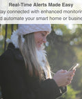 Person outdoors looking at smartphone for real-time alerts, surrounded by greenery, wearing a winter hat and coat. Text overlay about enhanced monitoring.