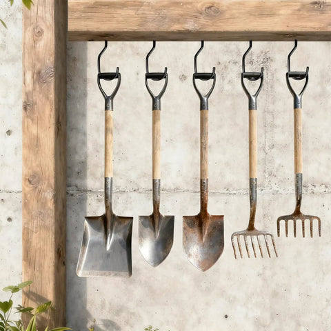 Four garden tools hanging on hooks, including shovels and a rake, against a wooden and concrete background in outdoor setting.