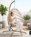 Woman relaxing in an indoor outdoor wicker hanging egg chair with stand cozy patio swing hammock egg shell chair with cushions 350 lb capacity sturdy porch swing for backyard balcony beige.