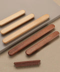 Two sets of long, slim wooden drawer handles in light and dark wood tones, laid out on a beige surface, showcasing minimalist design.