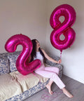 Woman holding hot pink number 2 and number 8 balloons on a couch, featuring Hot Pink Number 8 Balloon 40 Inch Giant Foil Balloon for Party Supplies.