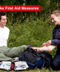 Front view of portable red travel first aid kit opened outdoors beside a backpack with medical supplies.