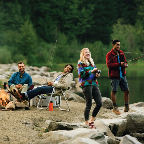 Group of people fishing by a lakeside with rods and a campfire, surrounded by rocks and trees.