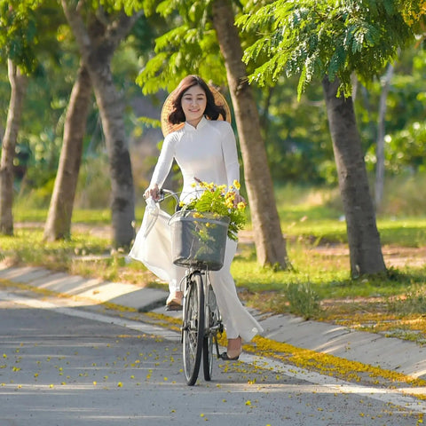 Person in white dress riding a bicycle on a tree-lined path with flowers in the basket, summer scene.