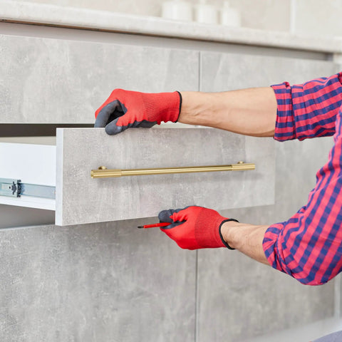 Man installing Top Tier Solid Brass Cabinet Pulls With Brushed Gold Finish on kitchen drawer with red gloves.
