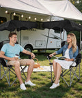 Two people sitting on Folding Camping Chair With Canopy Shade And Cup Holders, relaxing near RV with picnic setup.