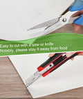 White ceramic fiber kiln paper sheet being cut with blue-handled scissors on wood.