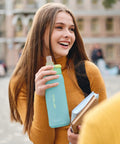 Young woman holding 24 Oz Insulated Stainless Steel Water Bottle With Straw Sky Blue outdoors, smiling with books.
