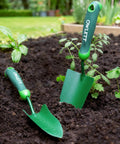 Two Go To Measuring Garden Trowels with ergonomic soft grip handles, one with a depth scale, in a garden bed among plants.