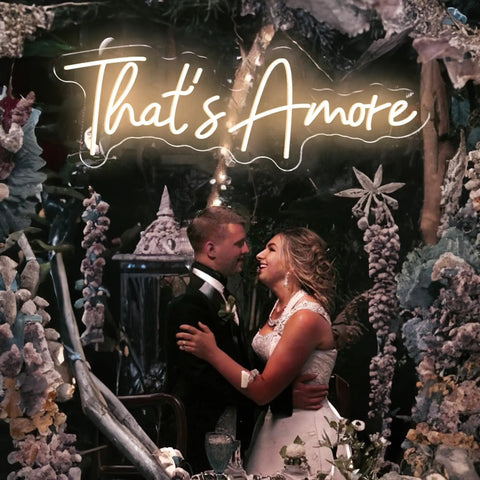 Couple under 'That's Amore' LED neon sign in wedding decor with flowers and lights.
