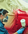 Person using spray gun to paint a car with a red finish while wearing safety gear and blue gloves.