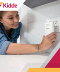 Woman installing carbon monoxide detector plugs into wall with AA battery backup and test hush button on a kitchen counter.