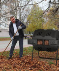 Dual Chamber Tumble Composter 43 Gallon Rotating Bin With 360 Degree Tumbler For Fast Easy Garden Waste Recycling BPA Free Black Outdoor Compost System next to a man raking leaves in a park.