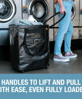 Person using 90L Collapsible Rolling Tote With Wheels For Groceries And Laundry in a laundromat, fully loaded, sturdy black design.